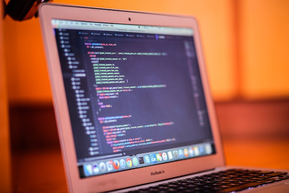 A close-up of a laptop screen displaying lines of colorful code, placed on a wooden desk with a warm, orange-toned background. The screen shows a dark-themed integrated development environment (IDE) w